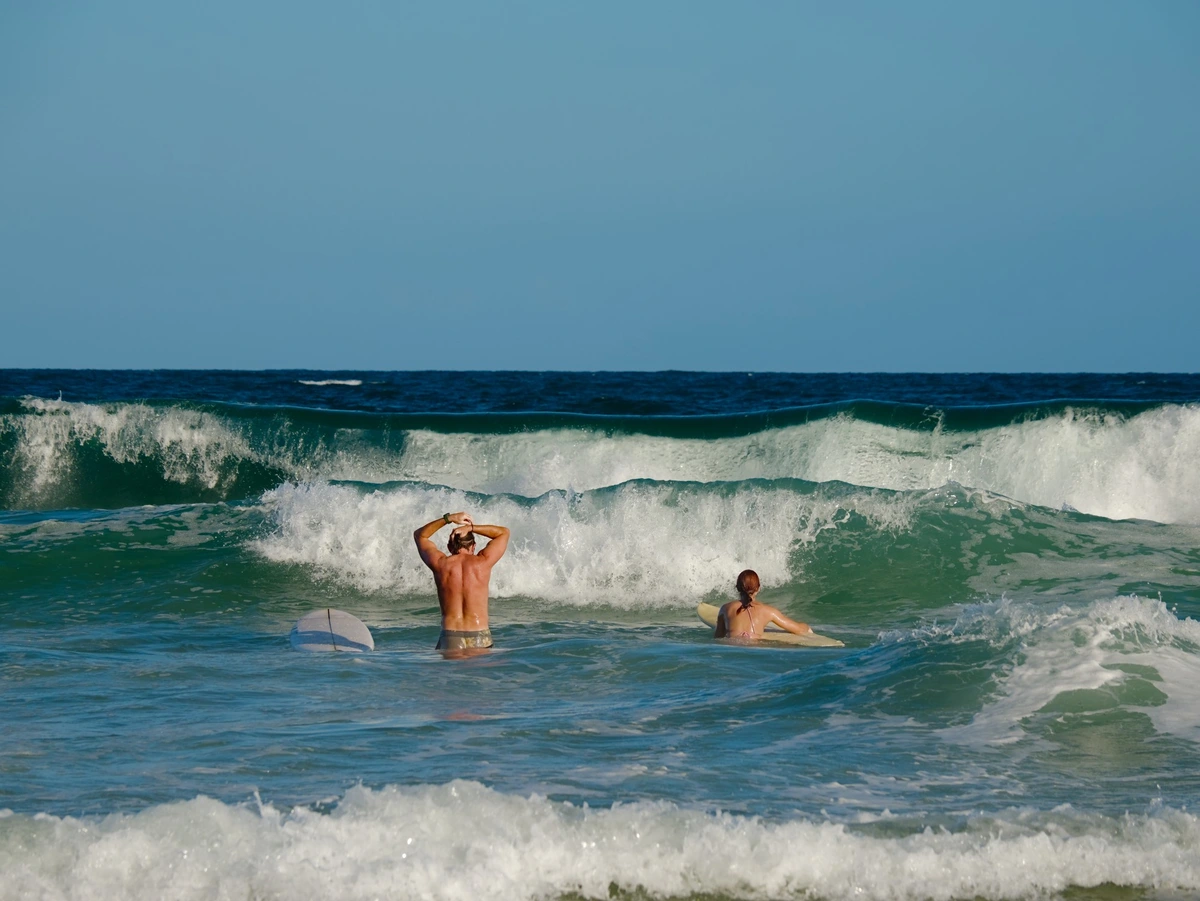 Two people with surfboardf standing in shallow surf facing the oncoming waves.  The man has his board beside him and is pulling his hair back in to a pony tail. The woman is crouched low, with her surfboard held close, as if she is about to climb on it.
