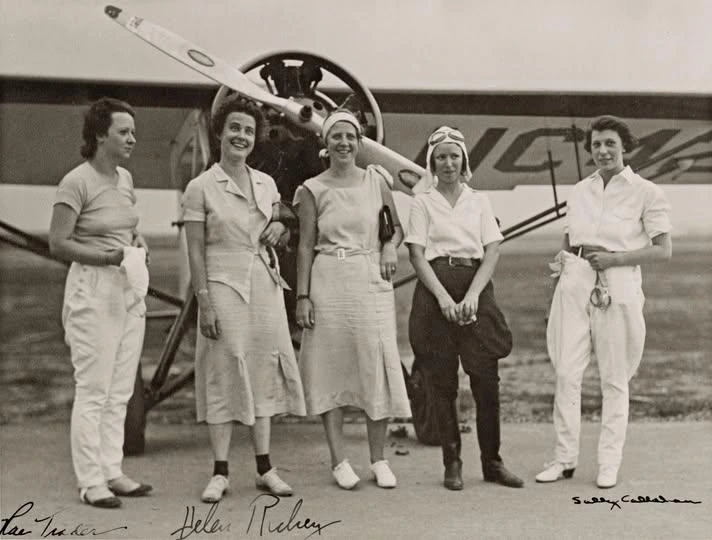 This black-and-white photograph shows five women aviators standing in front of a propeller aircraft, likely taken in the 1920s or 1930s during the golden age of early aviation. The women are dressed in a mix of practical and fashionable attire: two wear skirts and blouses, while the others sport trousers and flight gear, including one with a pilot’s cap and goggles.

They stand confidently, smiling or posing casually, exuding camaraderie and determination. Behind them, the airplane’s nose and large propeller dominate the background, reinforcing the pioneering spirit of the era.

At the bottom of the photo, some of their names are handwritten: Pan Tuckler, Helen Richey, and Sally Callahan, suggesting this image may have been a keepsake or publicity photo of notable female pilots.
