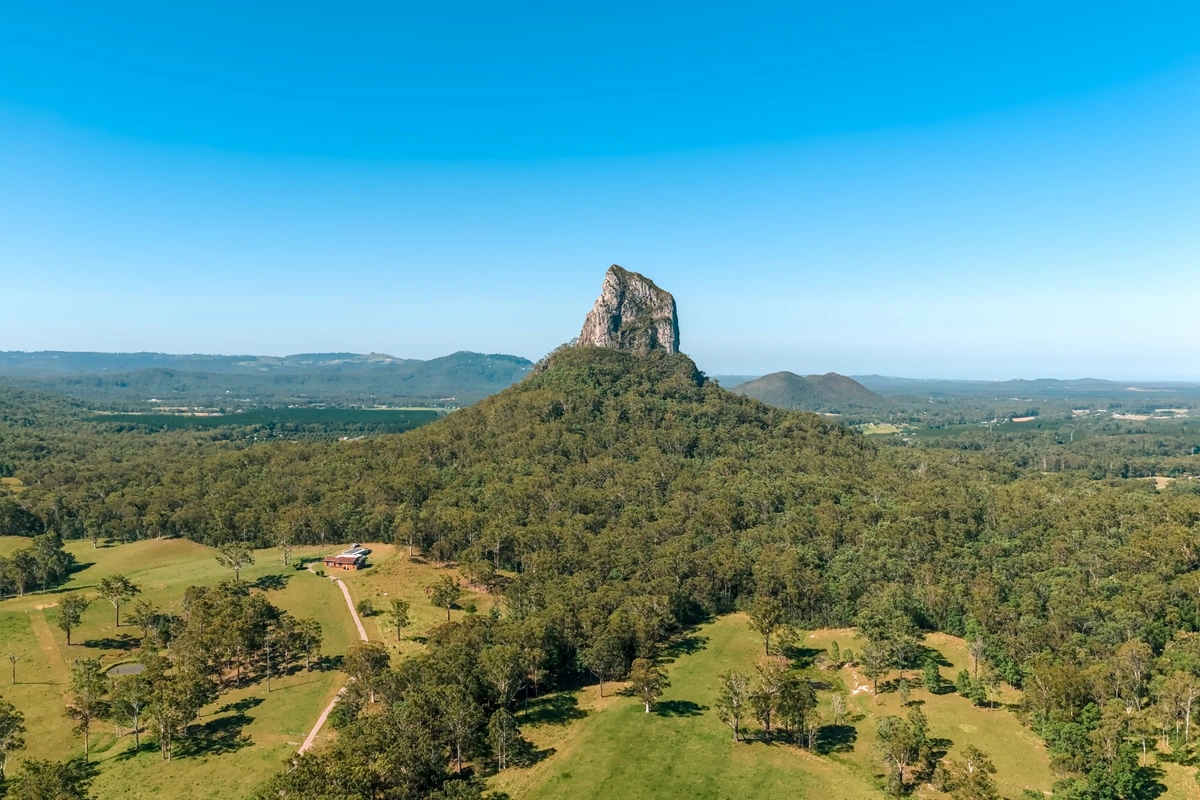 An aerial wide shot of the volcanic plug, Mount Coonowrin.  It's thrusts up out of a mostly flat, tree covered plain, with some small mountains visible in the distance.  A house surrounded by a wide grassy area is visible in on the plain in front of the mountain, standing out in an otherwise natural landscape